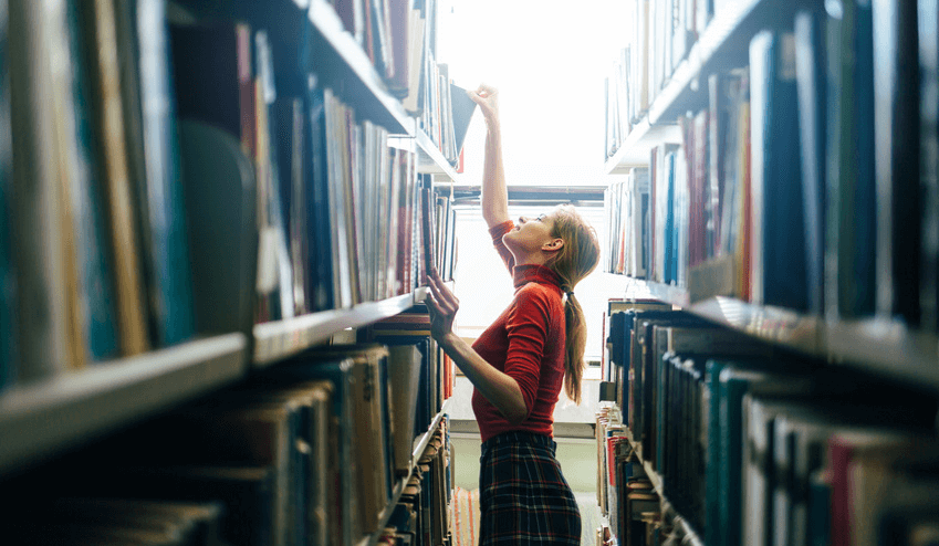 Young librarian searching books and taking one book from library bookshelf